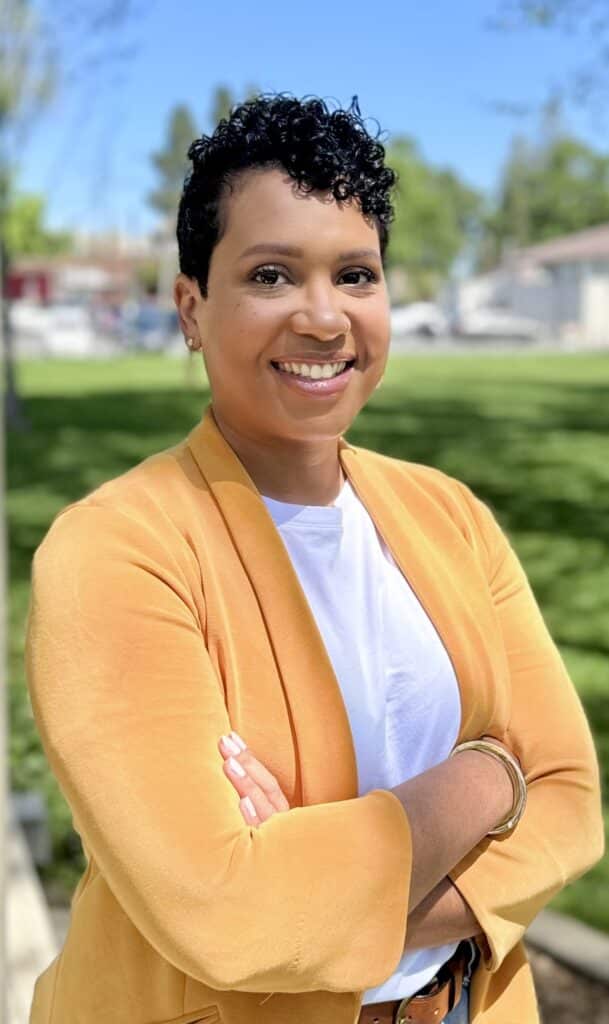 Smiling woman in a yellow blazer stands outside with arms crossed on a sunny day.