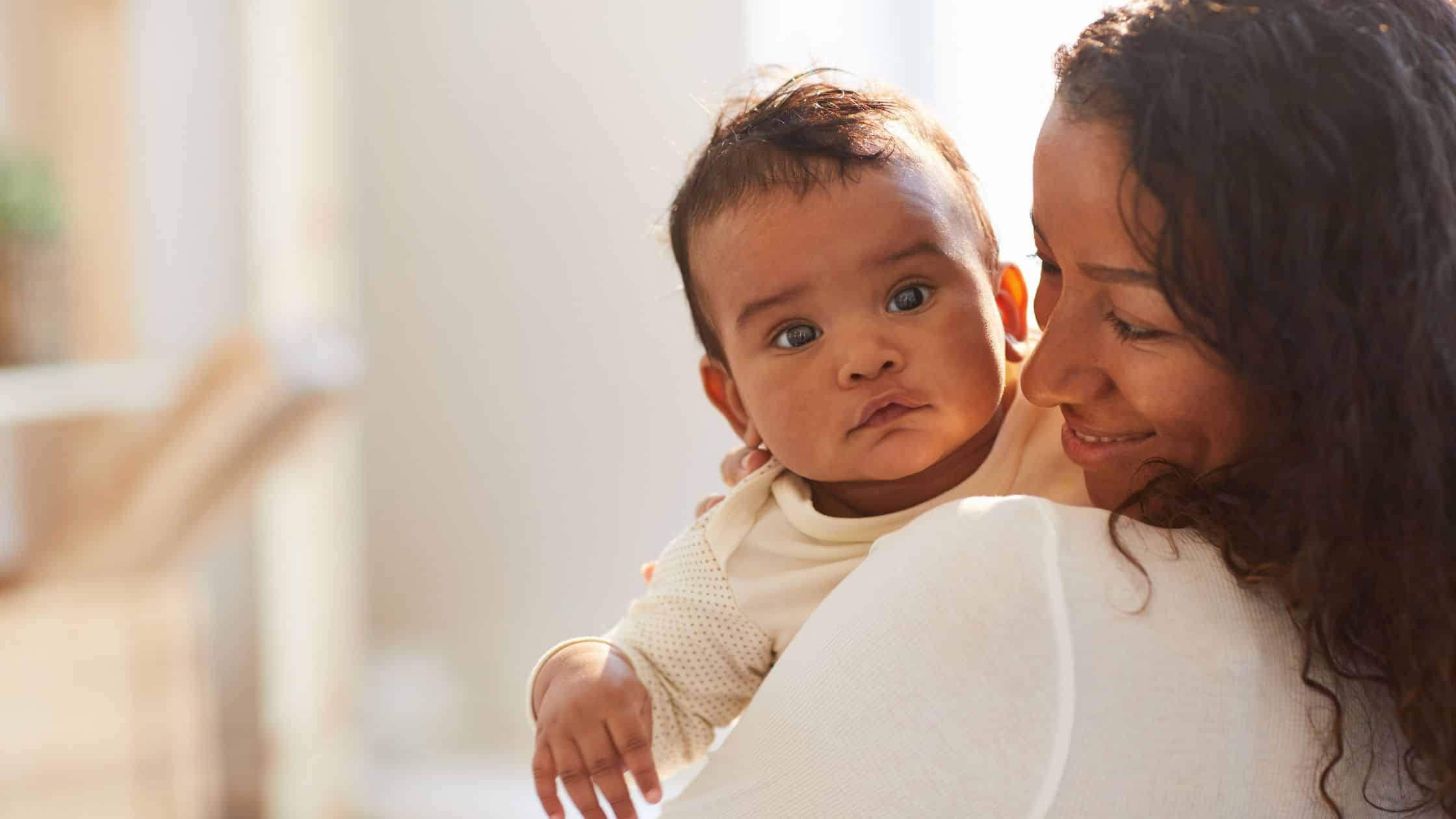 A mother holding her baby, who looks at the camera while she smiles lovingly.