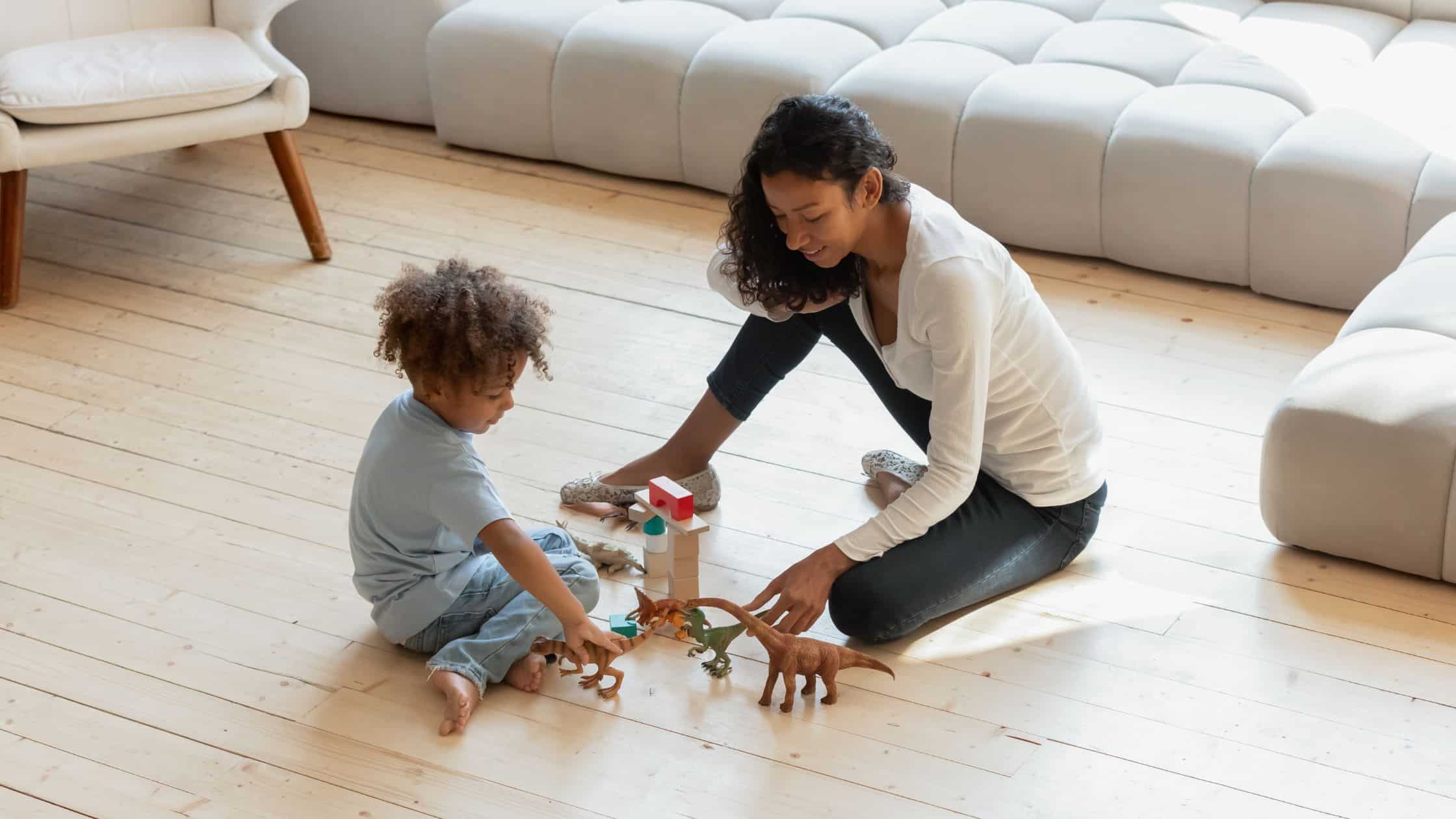 Woman and child sitting on the floor playing with toy dinosaurs and blocks in a bright living room, illustrating what a bilingual nanny for families does