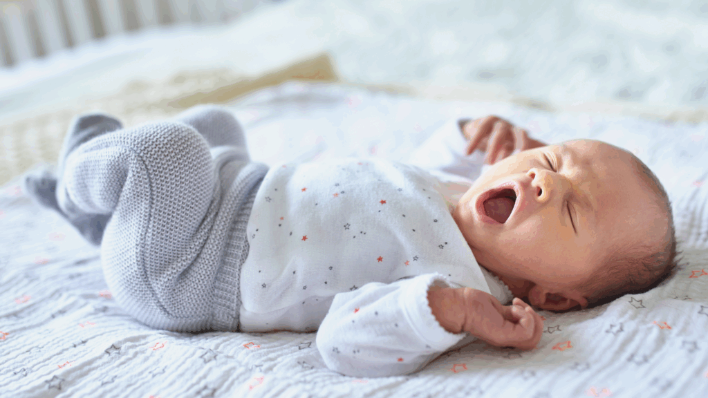 Newborn baby lying on a blanket, yawning in a white onesie and gray pants—perfectly peaceful, just like after care from a night nurse for newborns.
