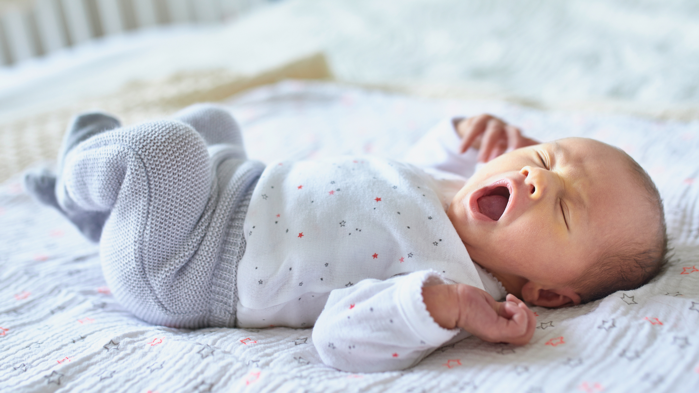 Newborn baby lying on a blanket, yawning in a white onesie and gray pants—perfectly peaceful, just like after care from a night nurse for newborns.