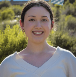 Young woman smiling outdoors with green bushes in the background, wearing a light-colored top.
