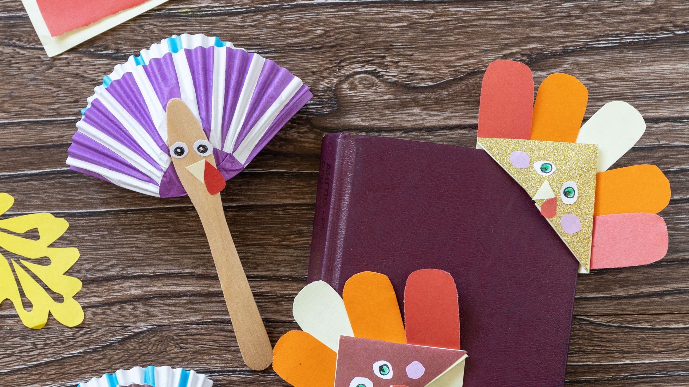 Colorful turkey crafts made from paper, cupcake liners, and popsicle sticks on a wooden table with a book—perfect Thanksgiving activities for preschoolers.