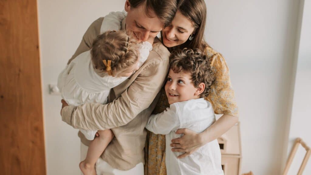 A smiling family of four hugs closely, with two parents holding their young daughter and son indoors.