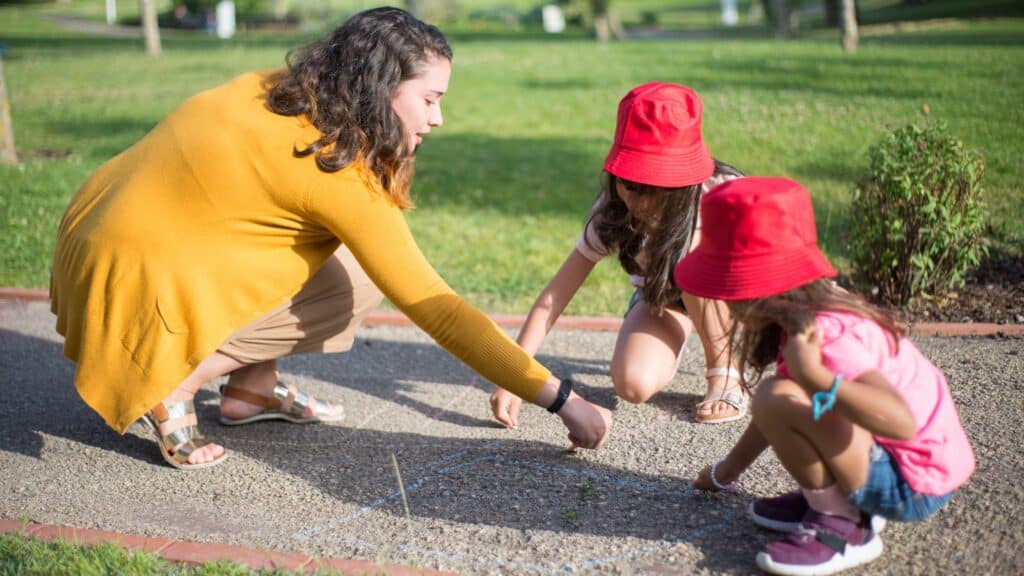 Woman and two children drawing with chalk on a path outdoors; both children wear red hats.