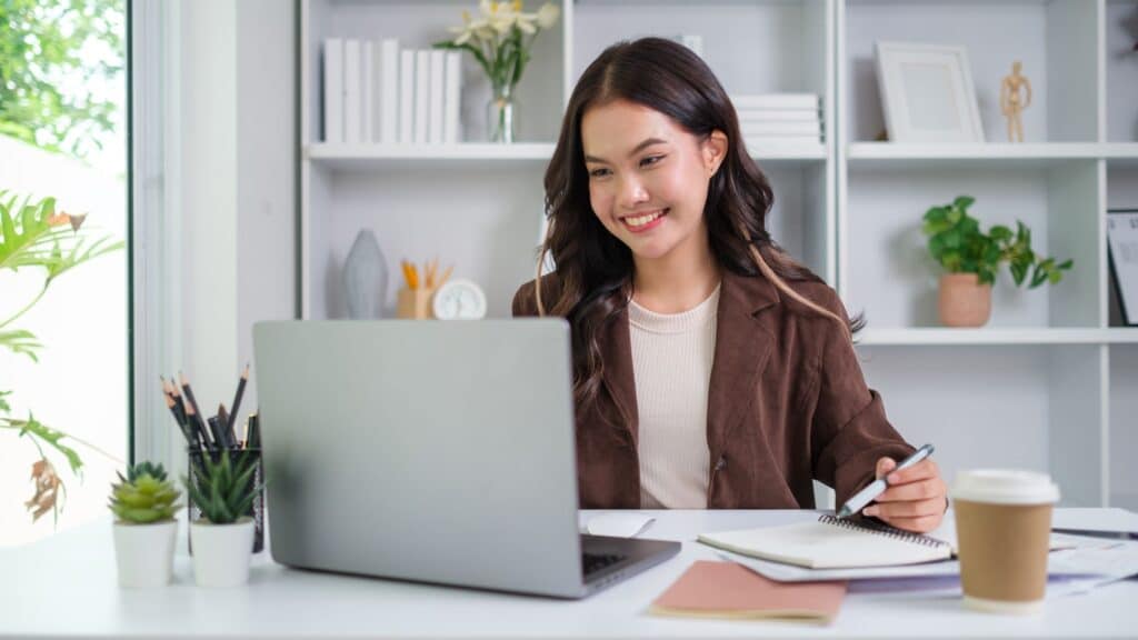 Woman smiling and working on a laptop at a desk with notebooks, pens, and a coffee cup in an office.