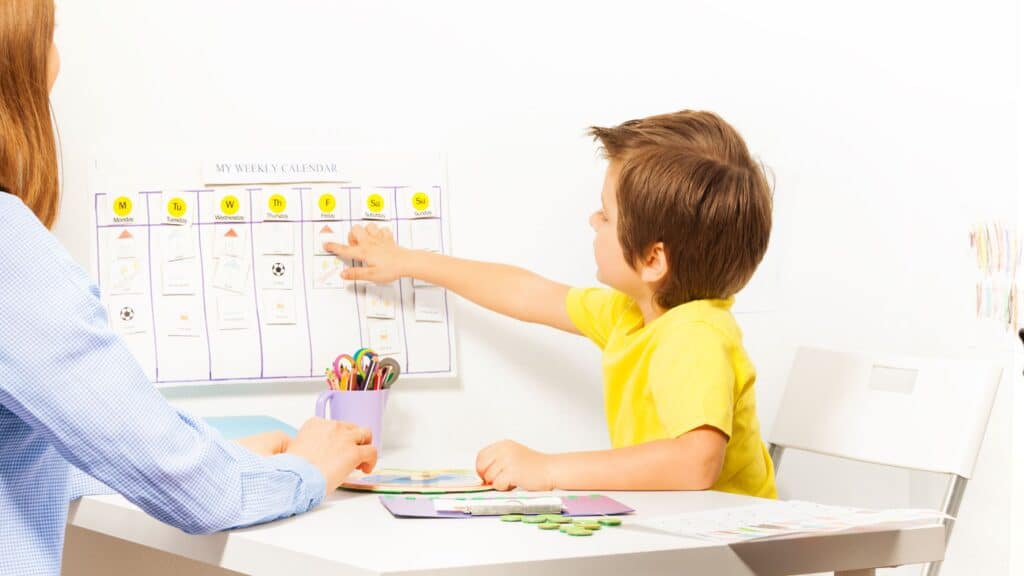 A boy points to a weekly calendar on the wall while sitting at a table with an adult, illustrating effective household system organization.