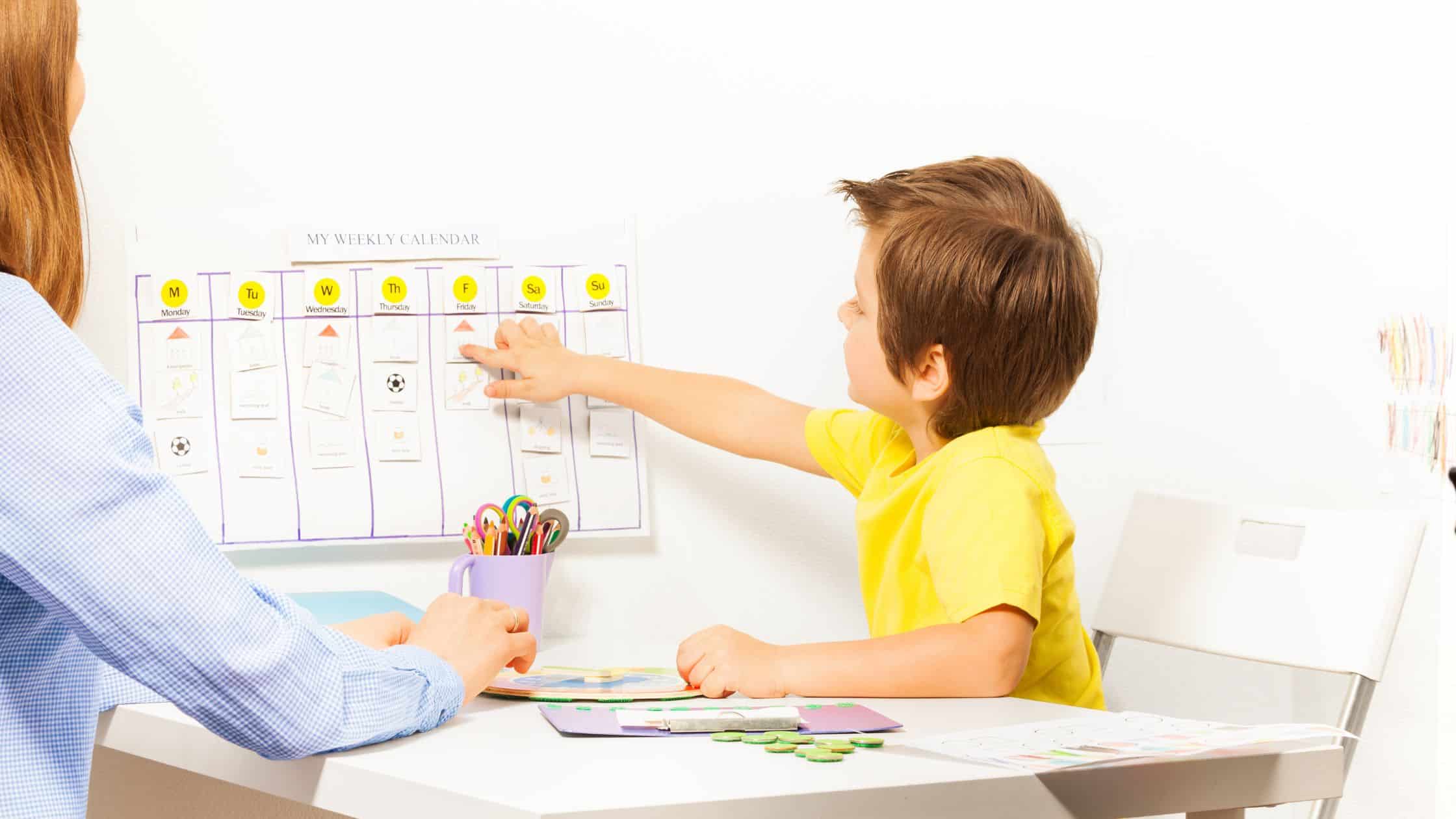 A boy points to a weekly calendar on the wall while sitting at a table with an adult, illustrating effective household system organization.