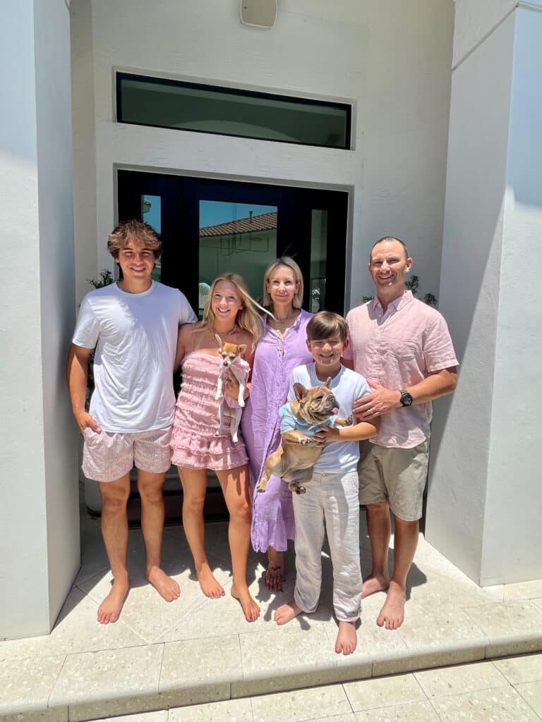 A family of five with two small dogs stands barefoot in front of a house, smiling on a sunny day, just after their Keesha Scott interview.
