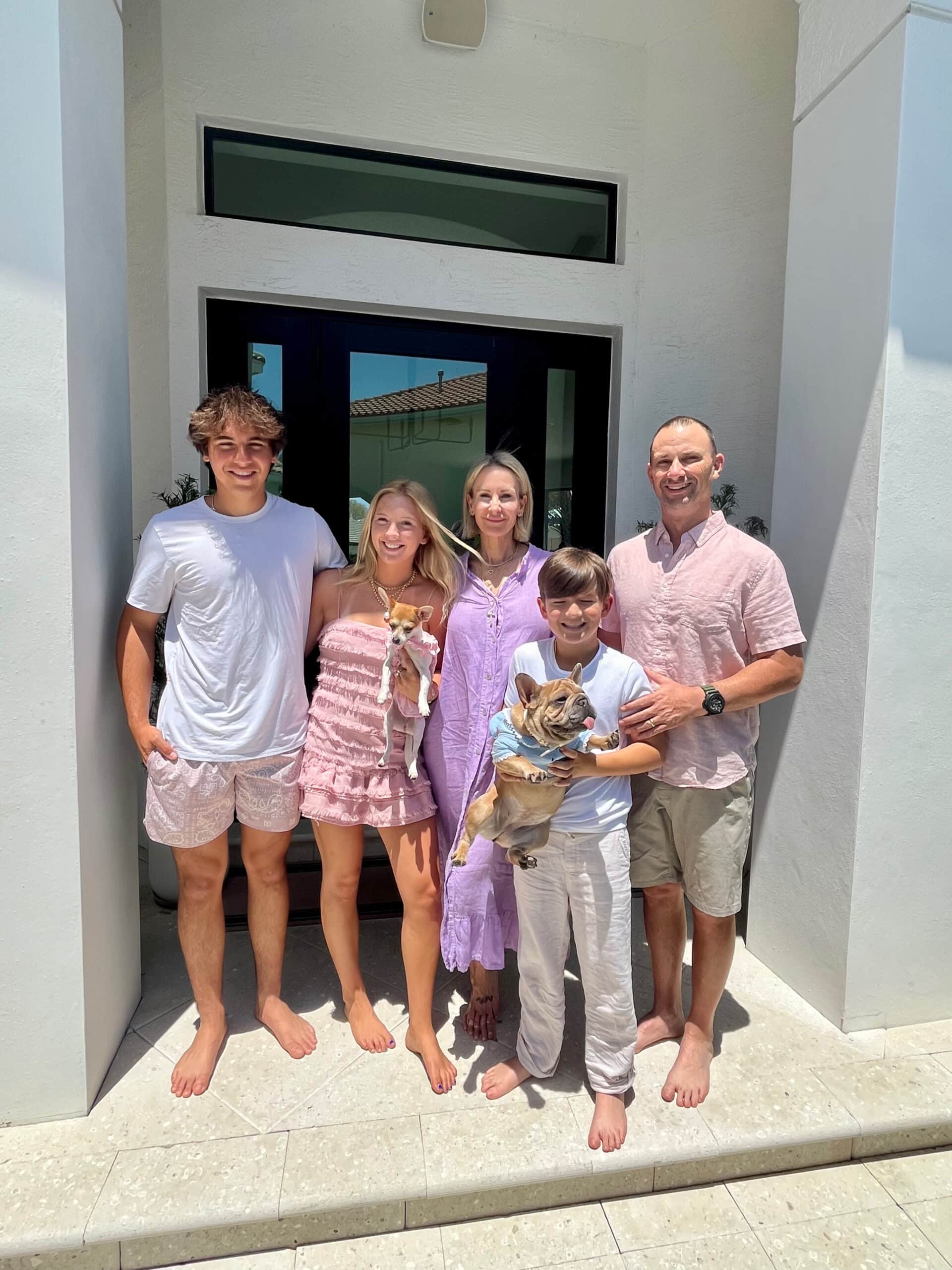 A family of five with two small dogs stands barefoot in front of a house, smiling on a sunny day, just after their Keesha Scott interview.