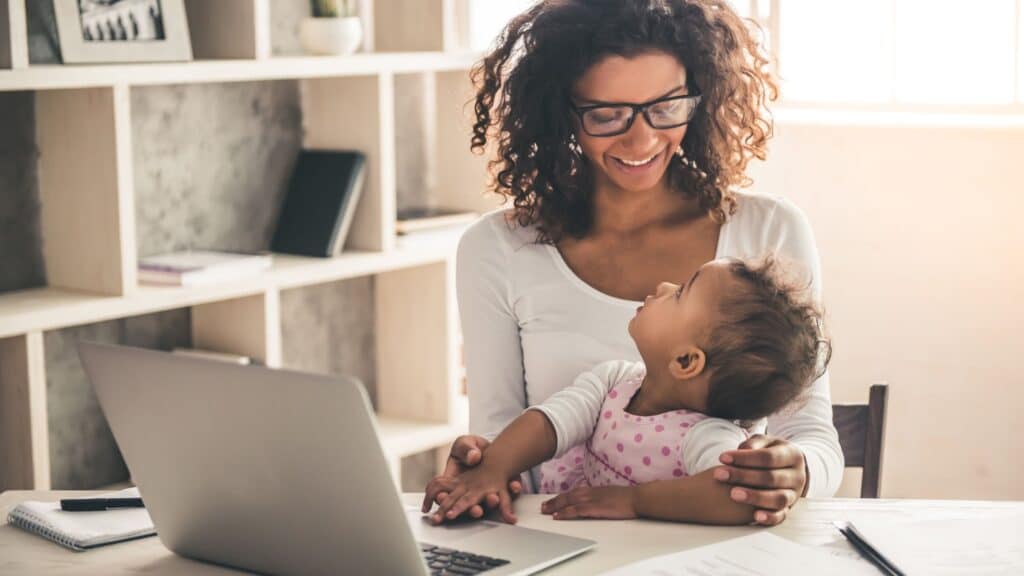 Smiling woman works on a laptop while holding a baby on her lap in a home office, showing the importance of protecting moms’ mental health while balancing work and family.