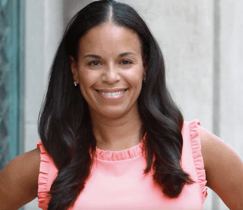 A woman with long dark hair smiles, wearing a sleeveless coral top, standing in front of a light-colored wall.