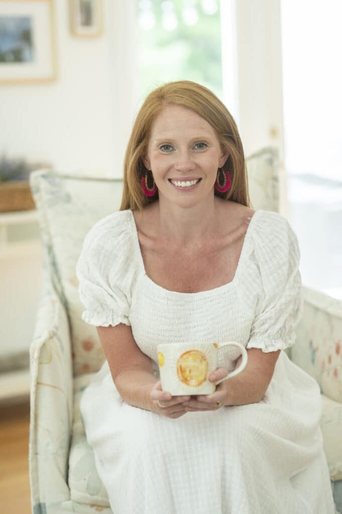 A woman in a white dress sits on a chair, smiling and holding a mug with both hands.