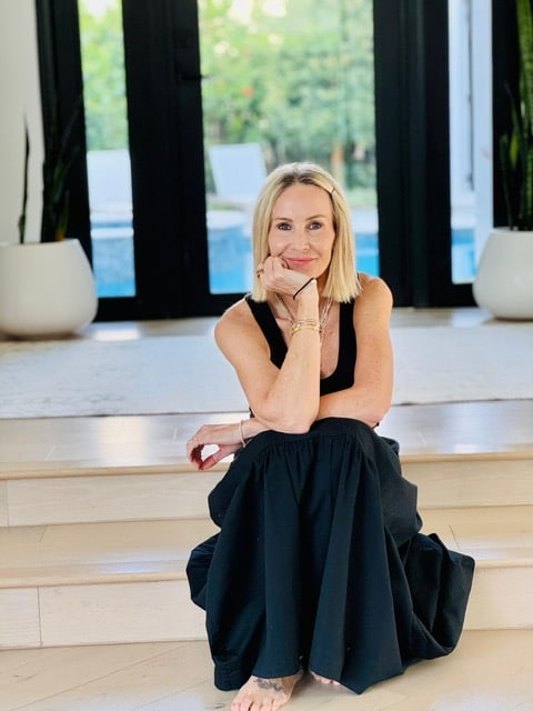 Woman with blonde hair in black dress sits barefoot on indoor steps, smiling with hand on chin.