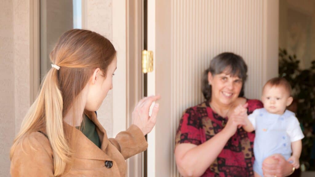 Woman waves goodbye at a door to a smiling older woman holding a baby inside, highlighting the importance of valuing caregiving across generations.