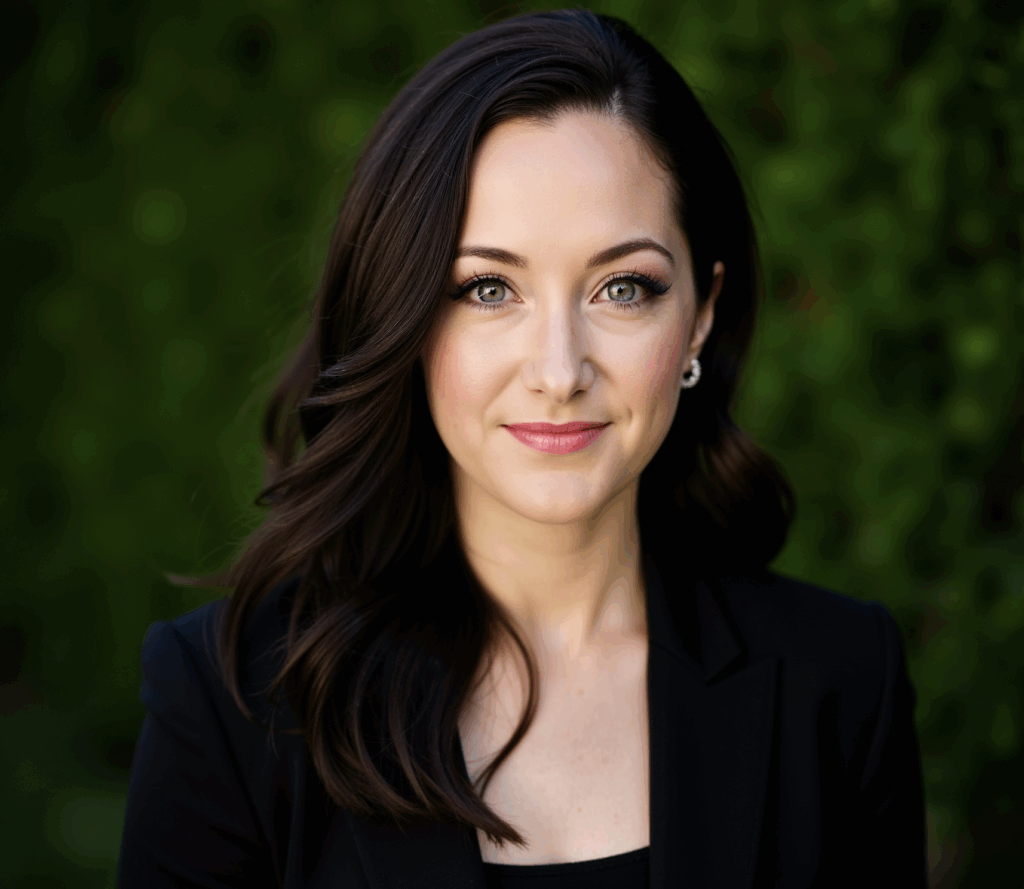 Woman with long brown hair in a black blazer, smiling softly, standing outside with green foliage in background.