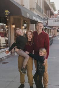 A smiling family of four poses together on a city sidewalk in front of shops and restaurants, reminiscent of a Chelsea Skaggs interview’s warm, candid atmosphere.