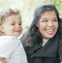 A woman and a young child smile together outdoors, both looking happy and cheerful.