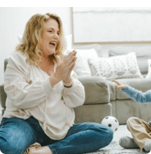 Woman clapping and smiling while sitting on the floor with a child in a cozy living room.